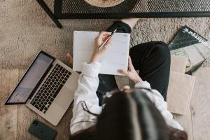 Woman sitting on the floor writing in a notebook with her laptop open